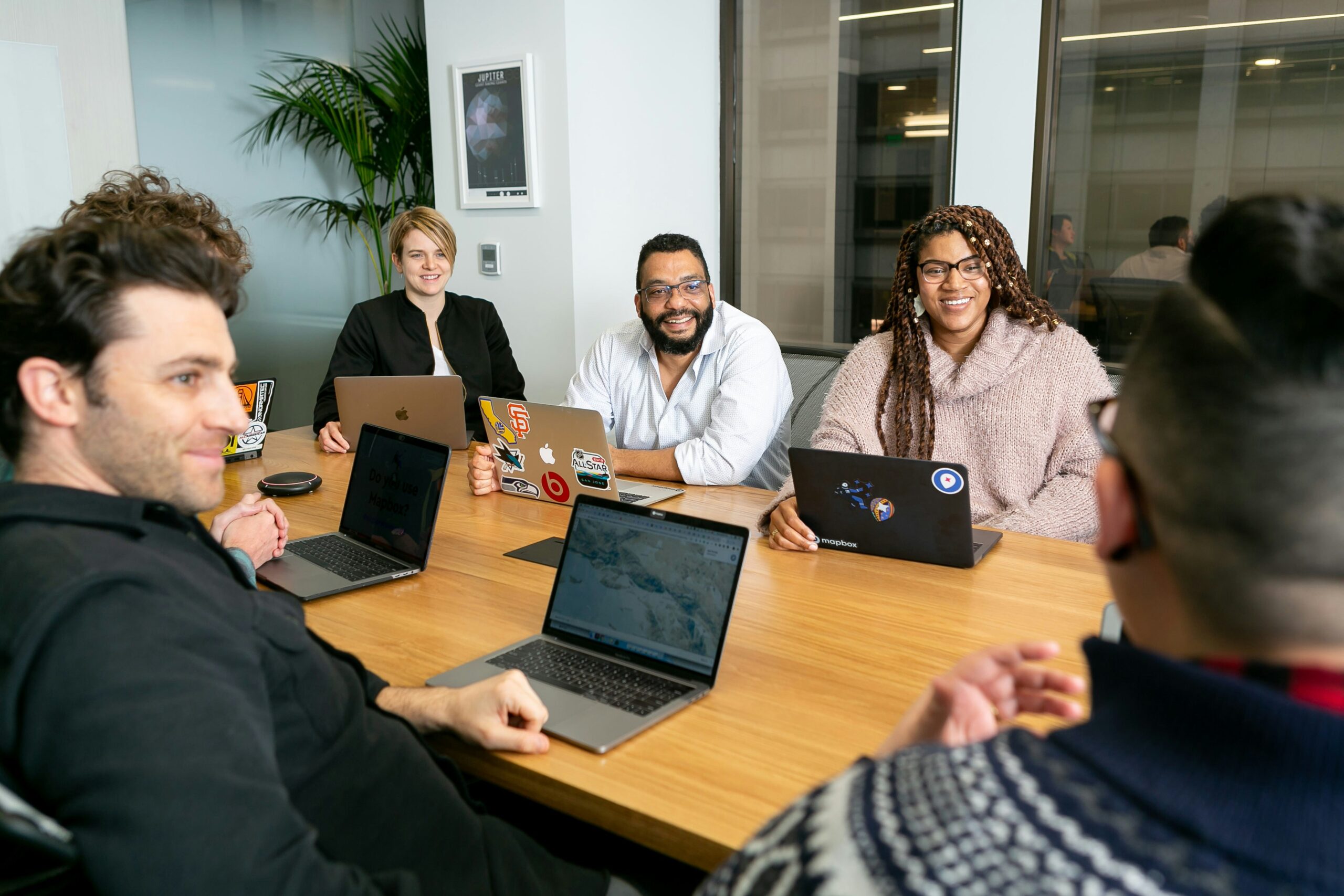 Six people sit around a conference table with laptops, smiling and talking during a meeting. The atmosphere appears relaxed and collaborative, with plant decor and large windows in the background.