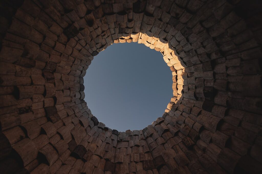 View from the bottom of a circular stone structure looking up at the blue sky through the opening, with sunlight illuminating part of the rough, stacked stones.