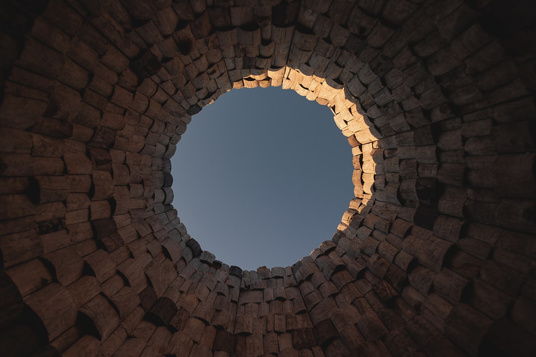 View from the bottom of a circular stone structure looking up at the blue sky through the opening, with sunlight illuminating part of the rough, stacked stones.